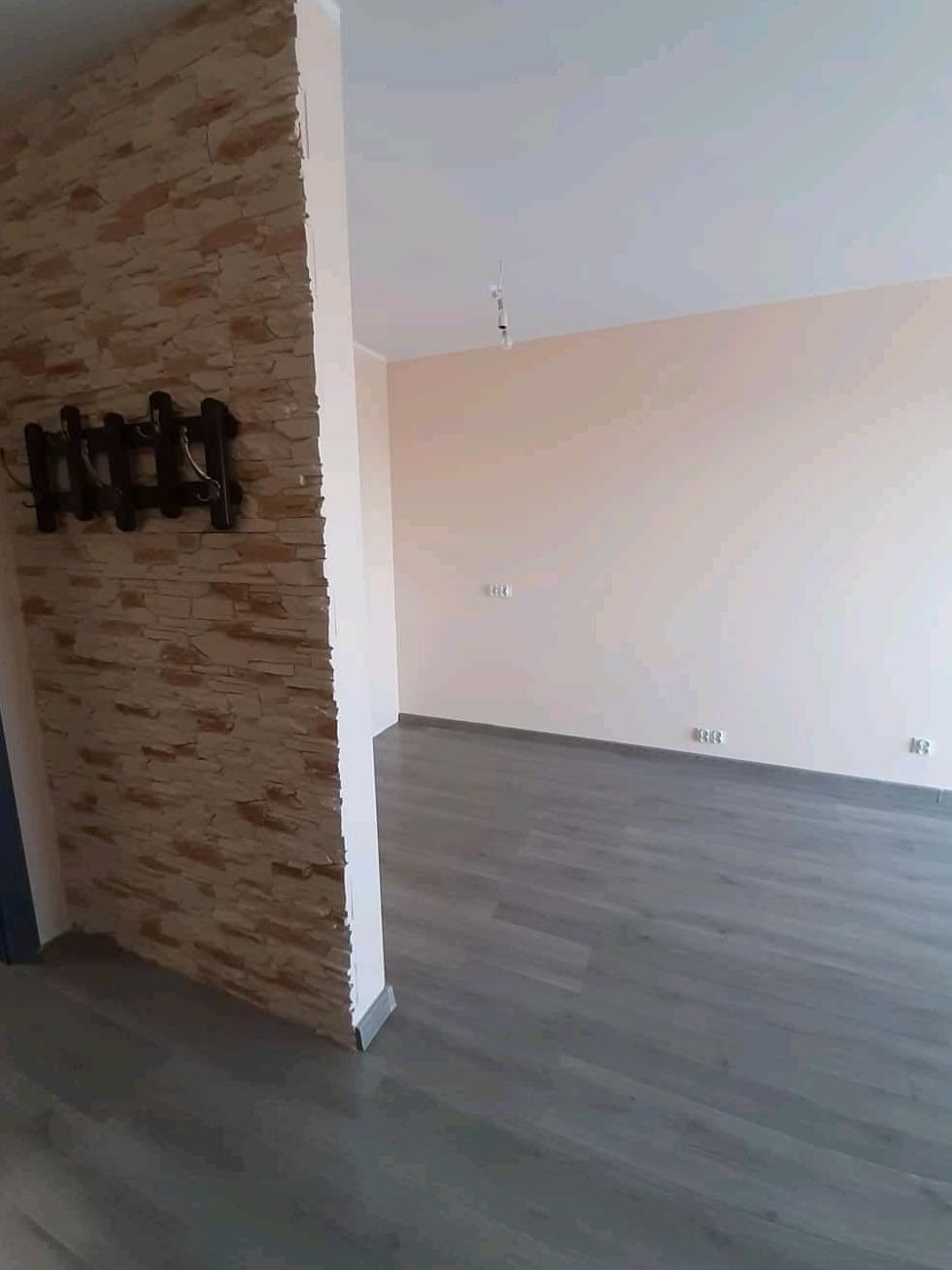 Świeżo wykończone wnętrze z dekoracyjną ścianą z imitacji kamienia, drewnianym wieszakiem na ubrania i szarą podłogą laminowaną, widok z perspektywy wejścia do pokoju.